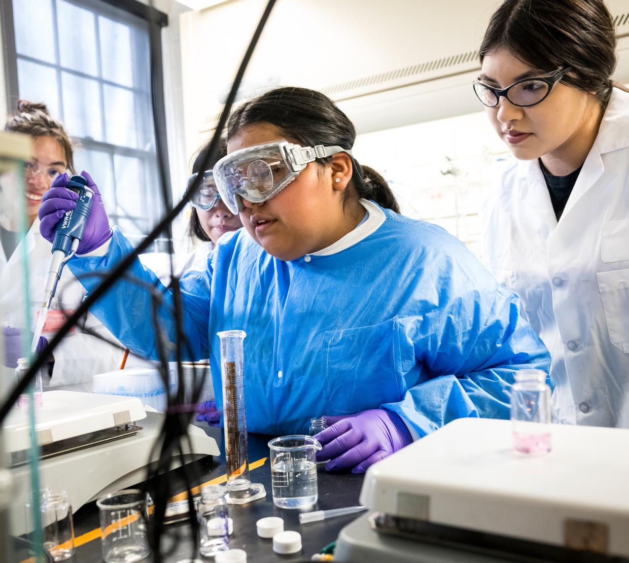 Students and staff clad in lab coats and glasses surround a lab table conducting experiments with pipettes, test tubes, beakers and other chemistry instruments in a lab at Oregon State University.