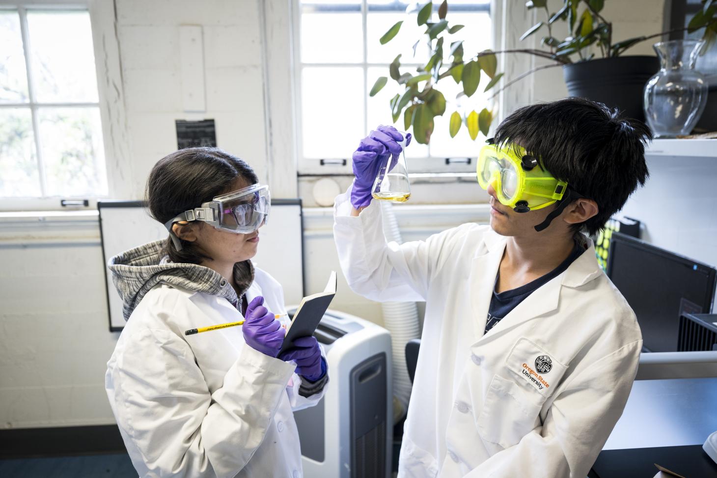 Two campers hold up a beaker with orange solution for a closer look and write down their observations at the Ignite inSTEM summer camp at Oregon State University.